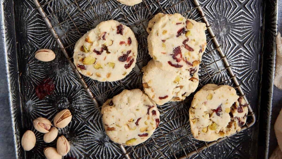 Five shortbread cookies with chopped pistachios and dried cranberries rest on a wire rack atop a textured baking tray. Some whole pistachios and dried cranberries are scattered nearby.