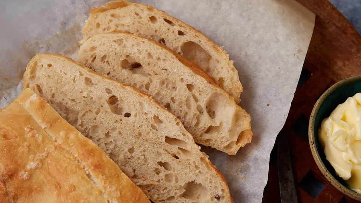 A loaf of rustic bread with a golden crust, partially sliced to reveal its airy crumb, sits on parchment paper atop a wooden board. A dish of butter and a knife are placed nearby on a blue surface.