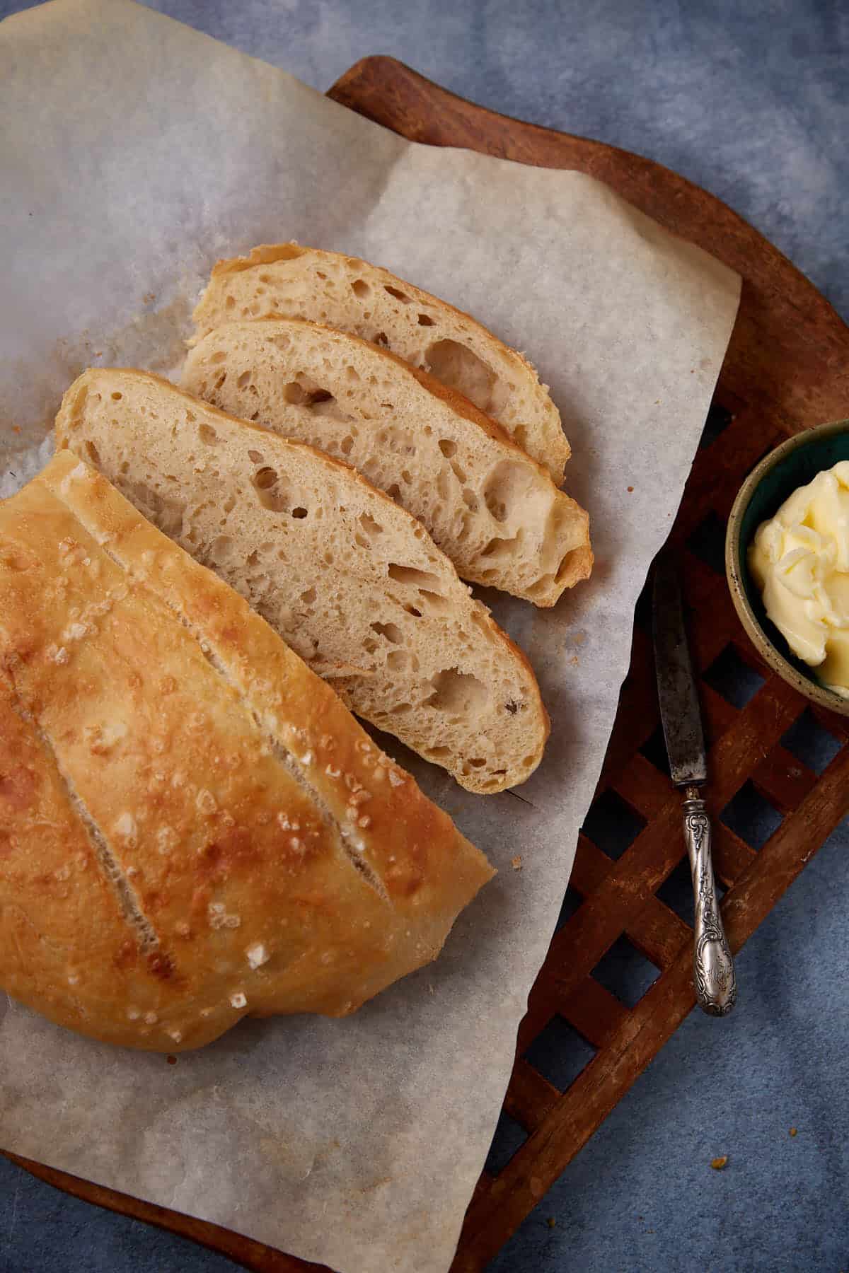 A loaf of rustic bread with a golden crust, partially sliced to reveal its airy crumb, sits on parchment paper atop a wooden board. A dish of butter and a knife are placed nearby on a blue surface.