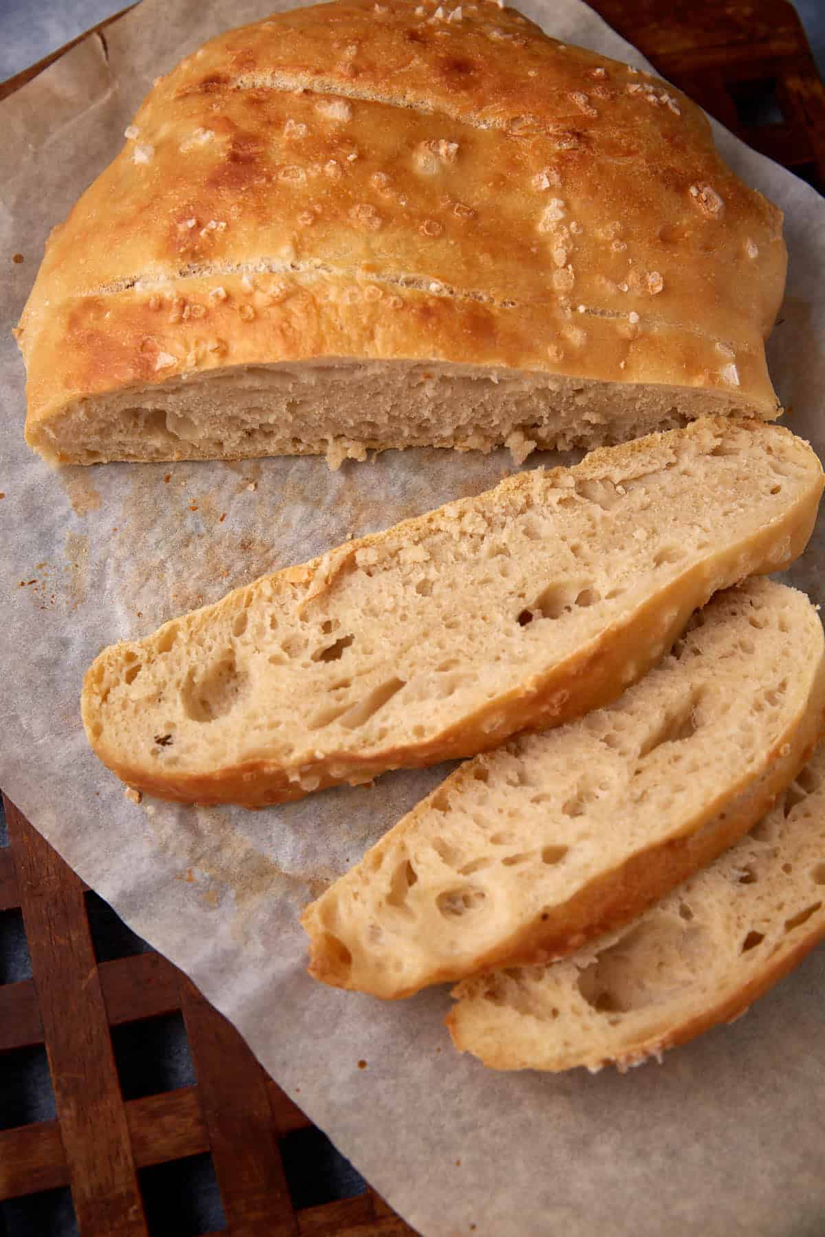 A round loaf of rustic bread with a golden crust, topped with coarse salt, sits on parchment paper. Three slices are cut and laid out in front of the loaf, showing a soft, airy interior with visible holes.