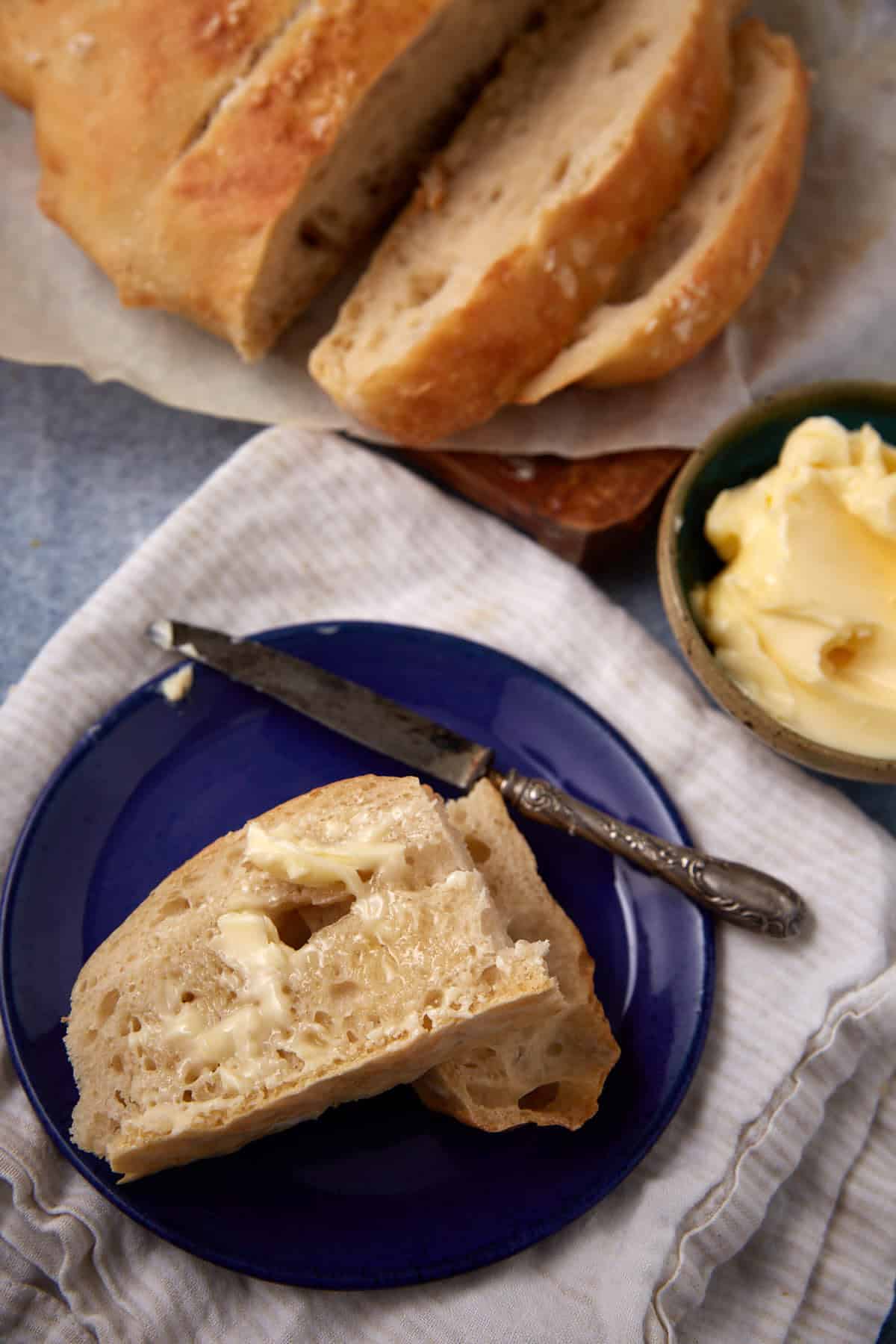 A slice of rustic bread with melting butter sits on a blue plate beside a vintage knife, with more sliced bread and a small bowl of butter nearby on a light cloth.