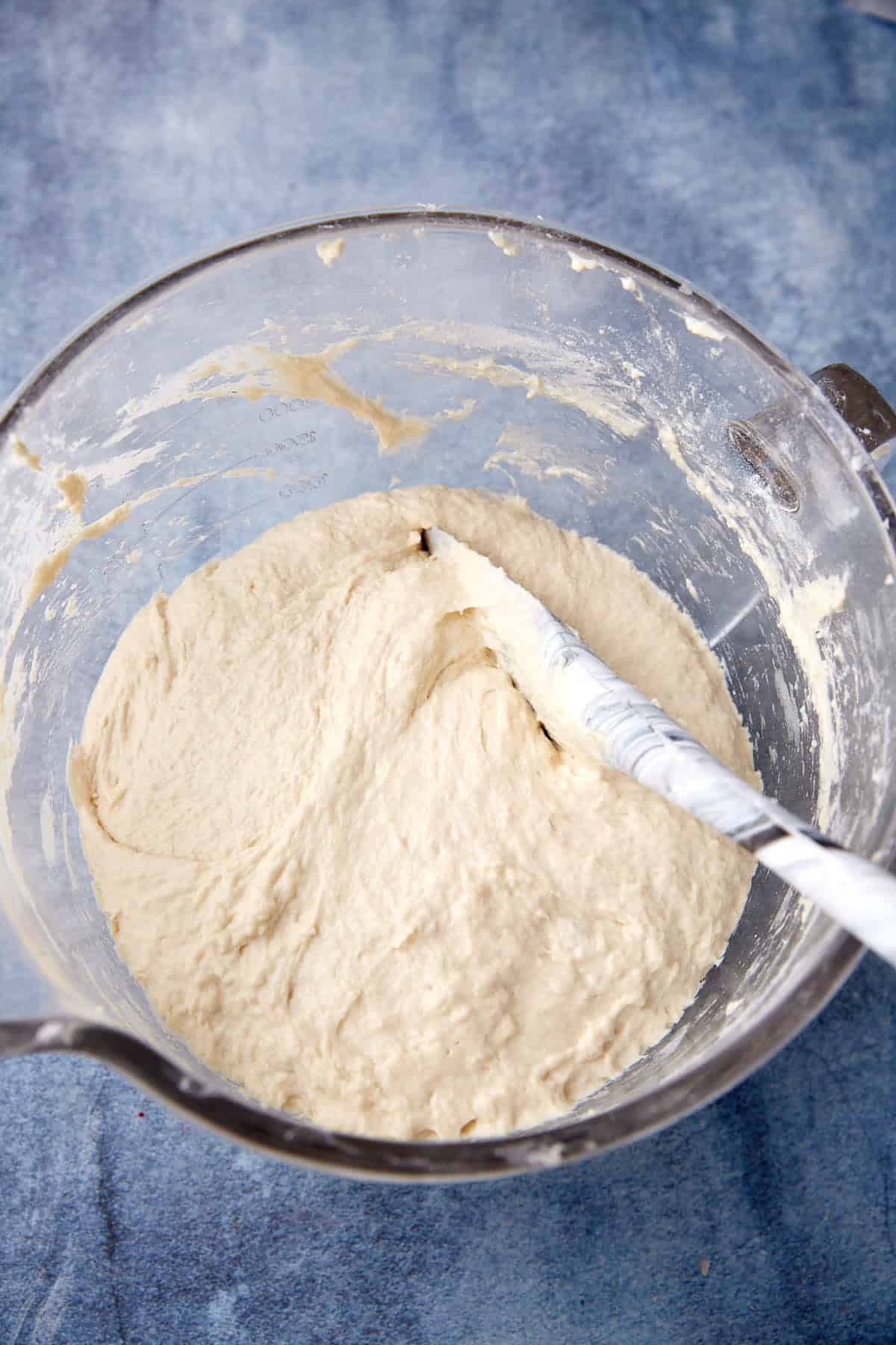A glass mixing bowl filled with sticky, unbaked bread dough. A white spatula is partially submerged in the dough, and the bowl rests on a blue textured surface.