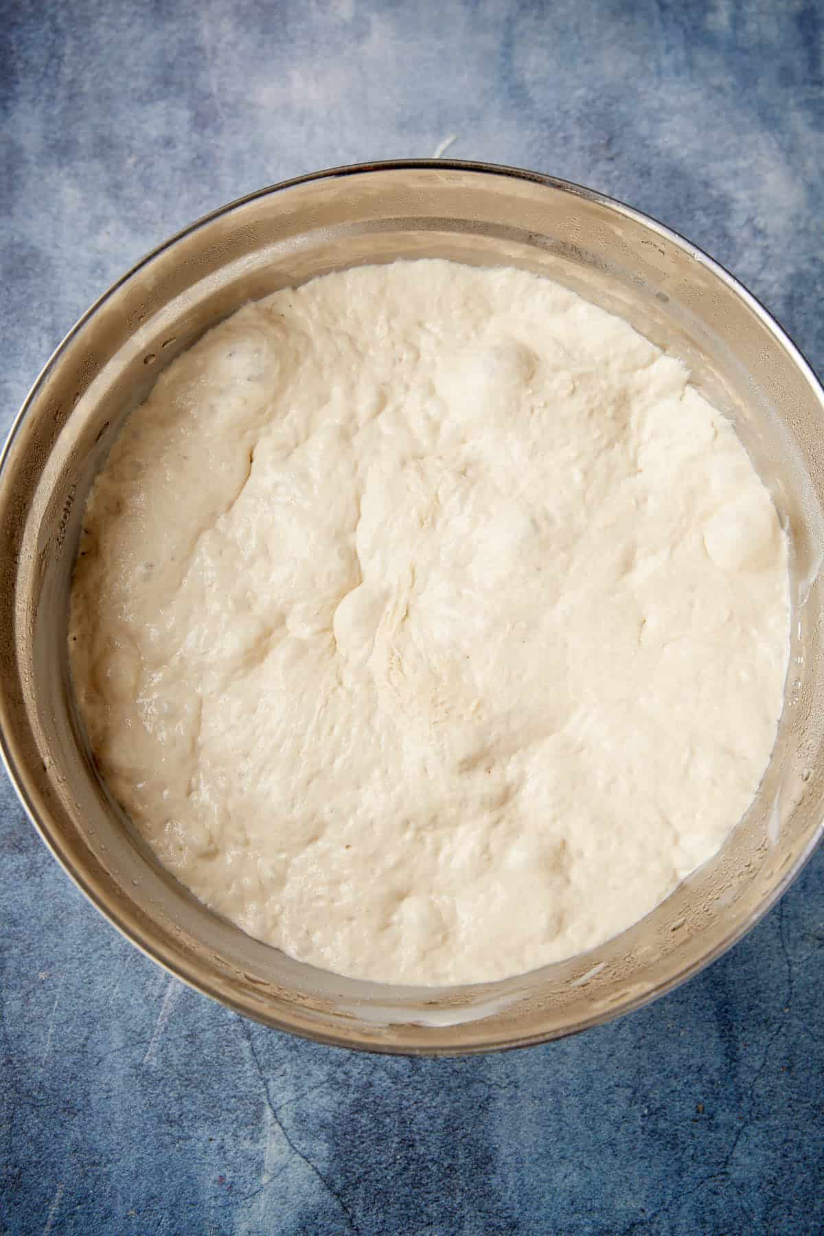 A metal mixing bowl filled with risen bread dough sits on a blue textured surface.