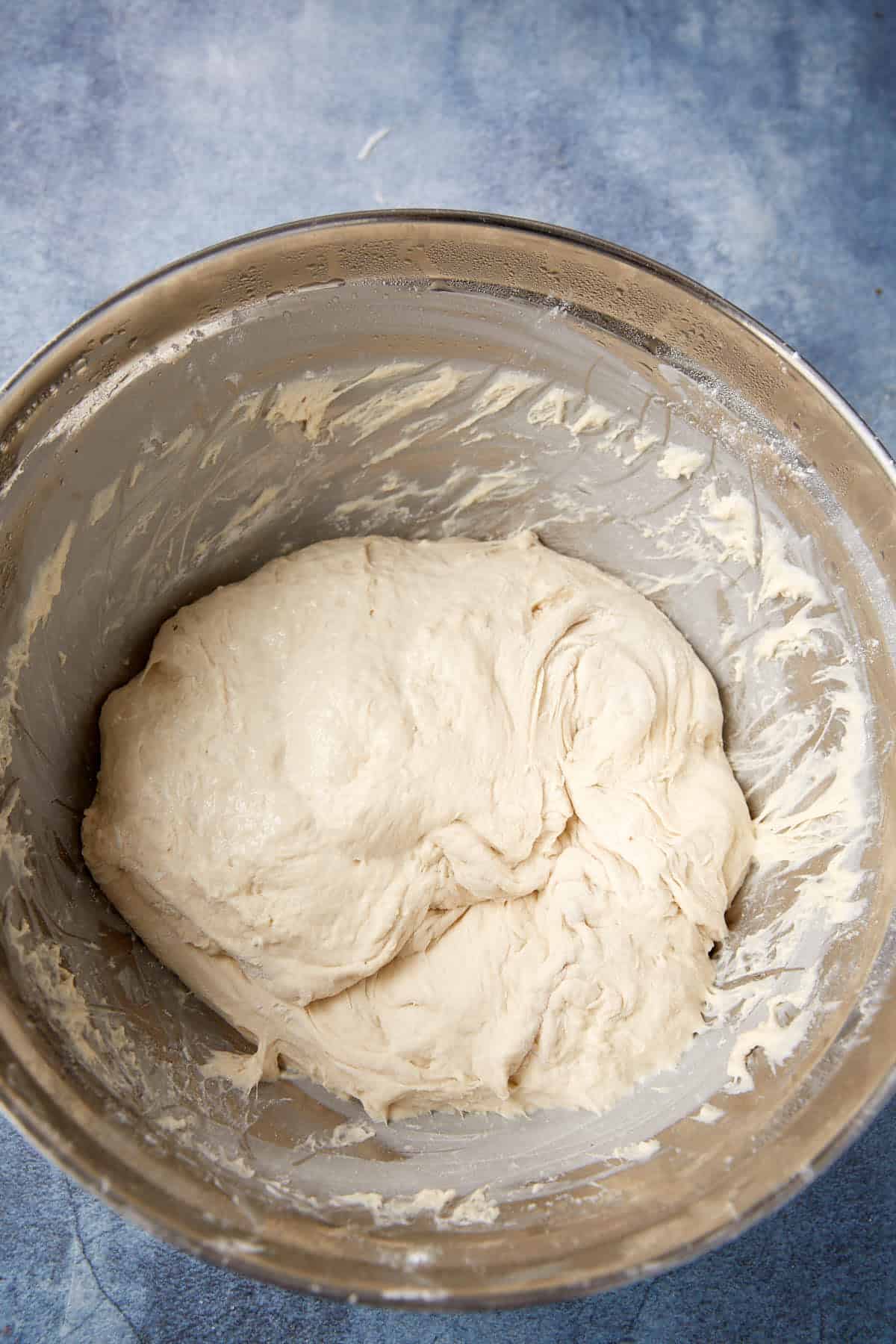 A stainless steel bowl containing a sticky, partially mixed bread dough sits on a blue countertop. The dough clings to the sides of the bowl, indicating it is in the early stages of preparation.
