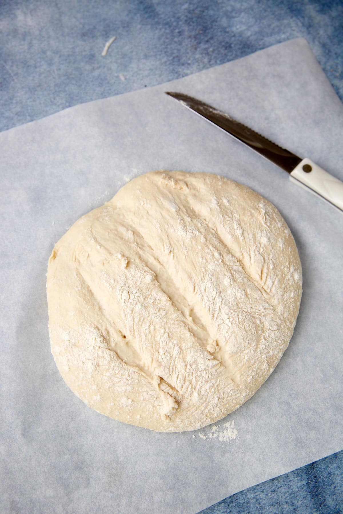 A round, unbaked loaf of bread dough with slashes on top sits on parchment paper. A white-handled knife is placed nearby on a blue surface.