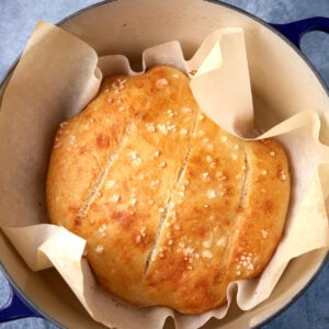 A round loaf of golden-brown bread with a crisp crust, sprinkled with flaky salt, sits on parchment paper inside a blue Dutch oven. Three shallow cuts are visible on top of the bread.