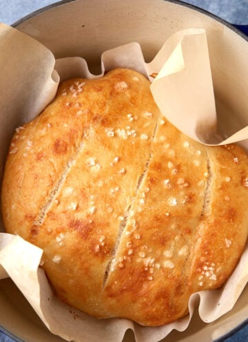 A round loaf of golden-brown bread with a crisp crust, sprinkled with flaky salt, sits on parchment paper inside a blue Dutch oven. Three shallow cuts are visible on top of the bread.