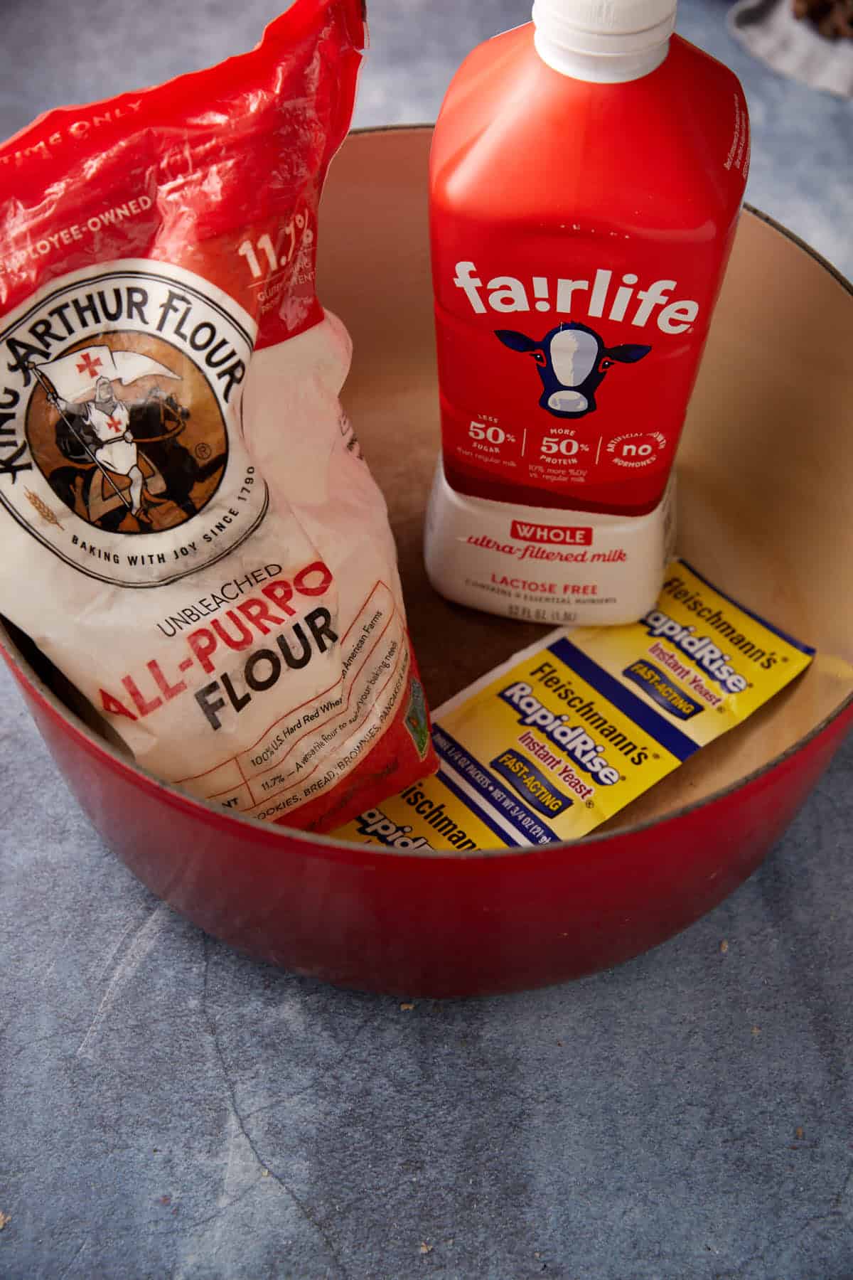 A red pot contains a bag of King Arthur all-purpose flour, a red carton of Fairlife whole milk, and three packets of Fleischmann’s RapidRise instant yeast on a blue countertop.