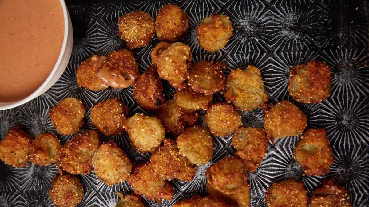 A batch of golden-brown Southern Fried Pickles is scattered on a dark textured tray, with a small bowl of creamy dipping sauce in the corner.