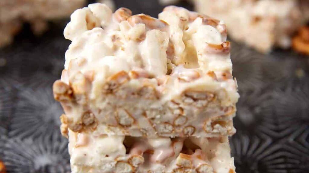 Two stacked white chocolate and pretzel bars with a creamy coating and visible pretzel pieces sit on a textured dark surface—no bake holiday desserts that look guest-ready.