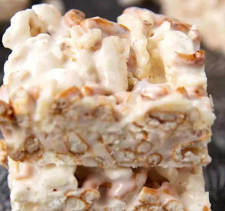 Two stacked white chocolate and pretzel bars with a creamy coating and visible pretzel pieces sit on a textured dark surface—no bake holiday desserts that look guest-ready.