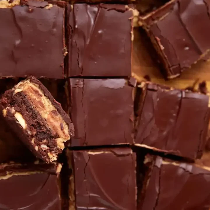 Squares of chocolate-topped dessert bars are arranged on a wooden surface. One bar is stacked on another, showing layers of chocolate, peanut butter, and a brownie-like base.