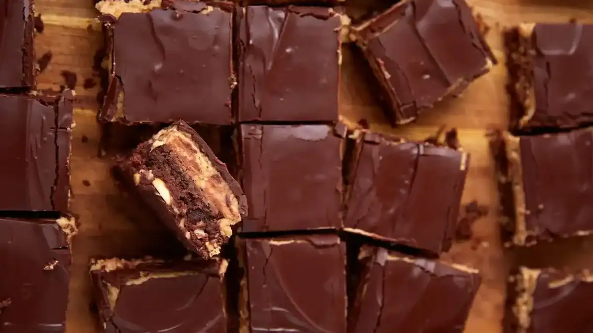 Squares of chocolate-topped dessert bars are arranged on a wooden surface. One bar is stacked on another, showing layers of chocolate, peanut butter, and a brownie-like base.