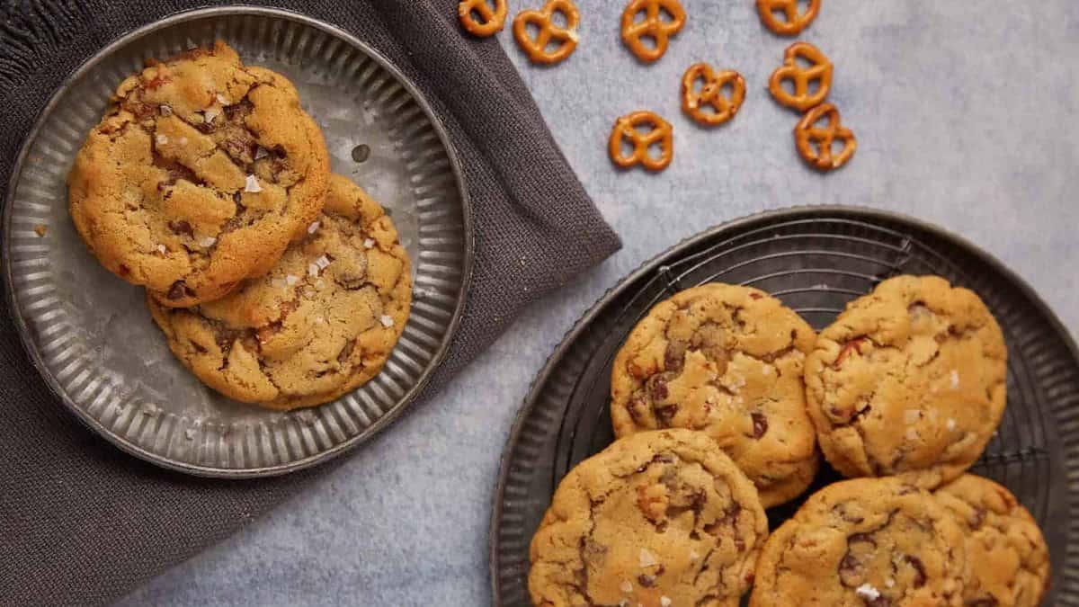 Two plates with chocolate chip cookies, one with two cookies on a gray cloth and the other with five cookies. Small pretzel pieces are scattered on the surface nearby.
