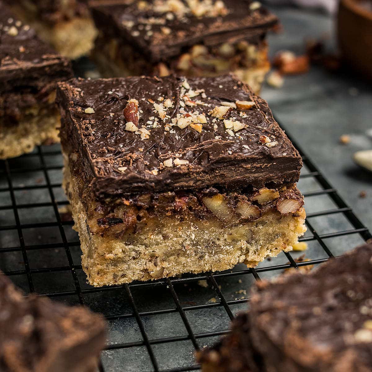 A close-up of a dessert bar with a thick, nutty base, chopped pecans, and a glossy chocolate topping rests on a black cooling rack—perfect alongside homemade Dutch Oven Bread.