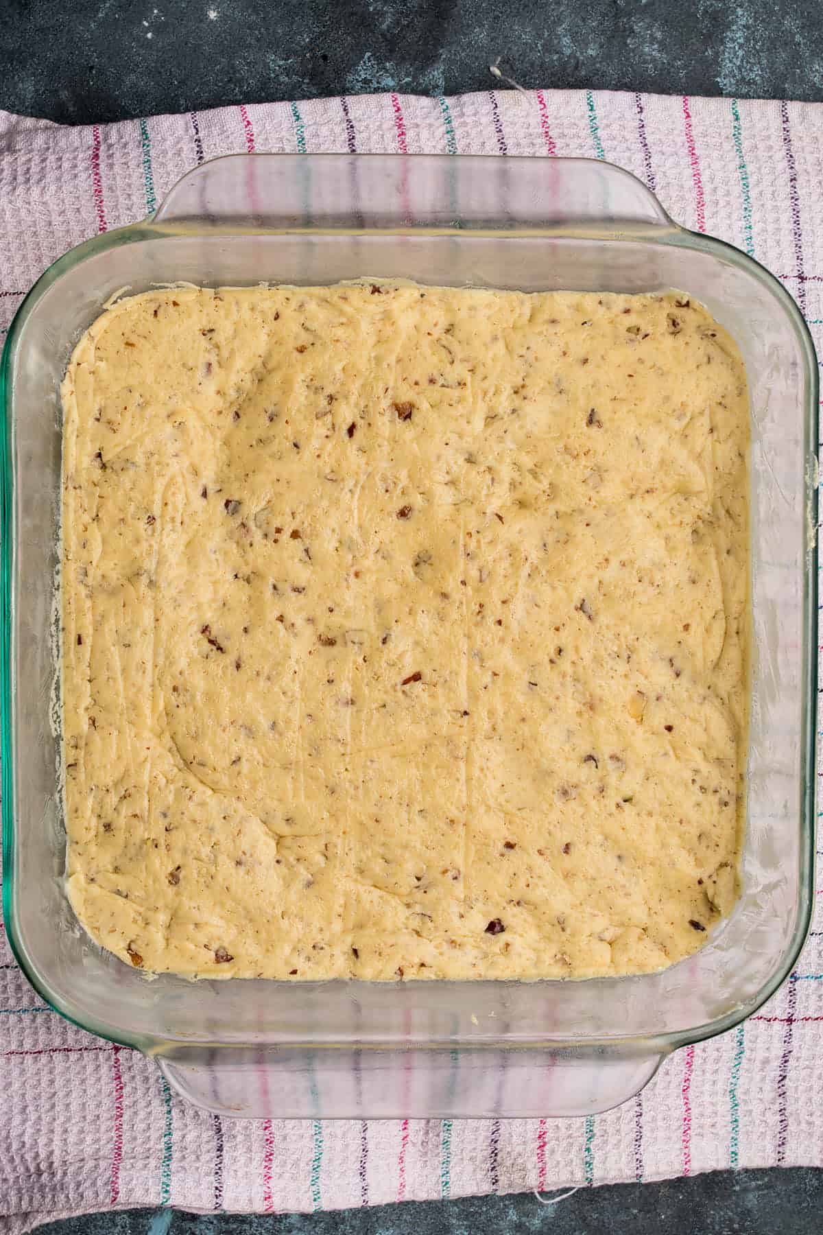 A glass baking dish filled with unbaked, textured cookie dough mixture, pressed evenly into the pan, rests on a striped kitchen towel—perfect next to a fresh loaf of Dutch Oven Bread.