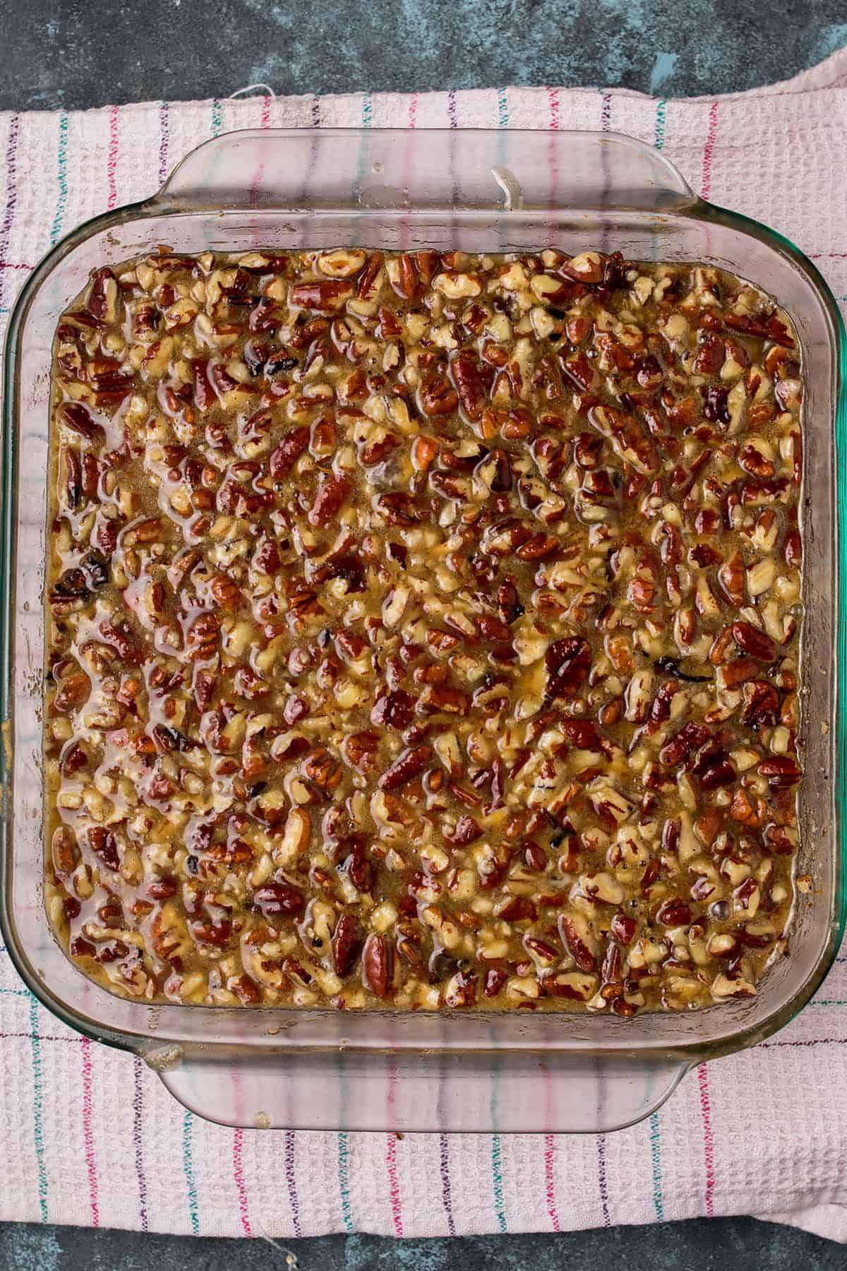 A glass baking dish filled with a pecan pie mixture topped with chopped pecans sits on a striped kitchen towel, ready to share space at the table with warm Dutch Oven Bread.