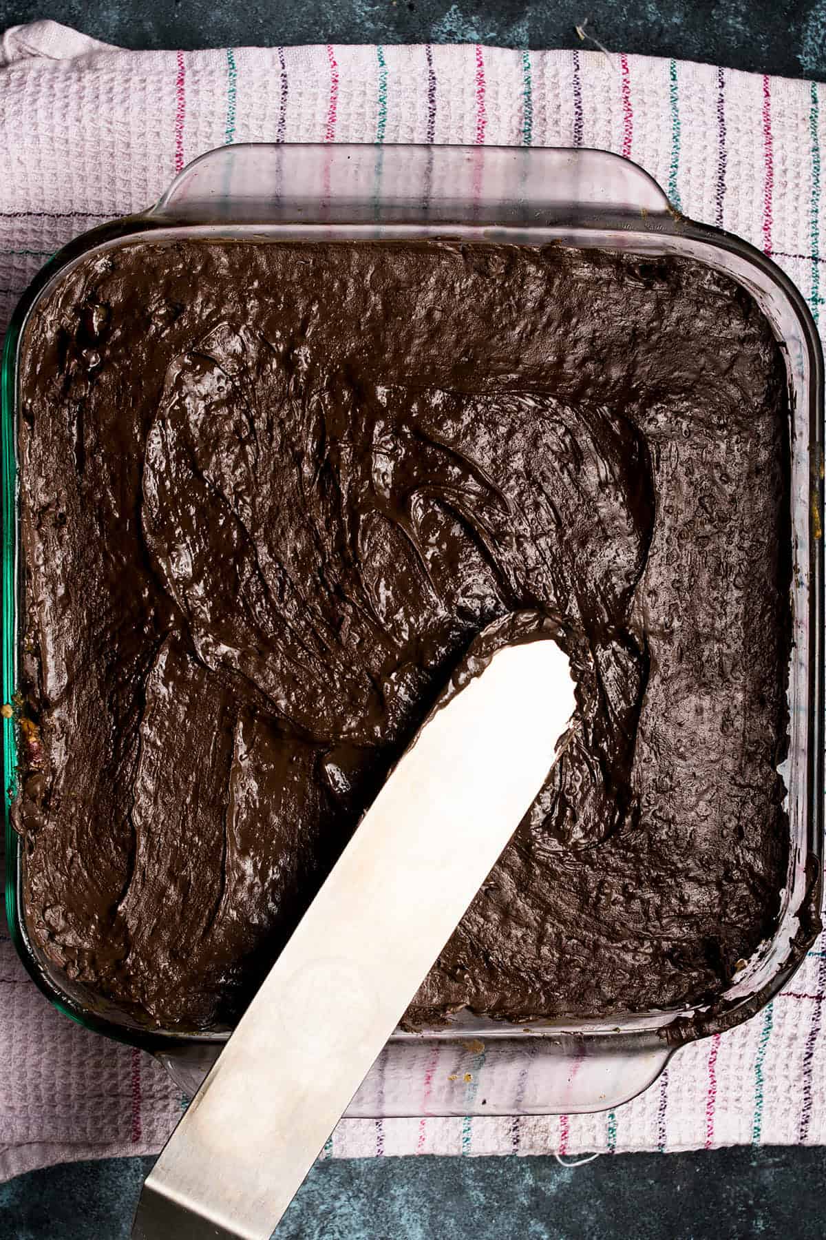 A spatula spreads chocolate frosting smoothly over a square cake in a glass baking dish, which sits on a striped kitchen towel next to freshly baked Dutch Oven Bread.