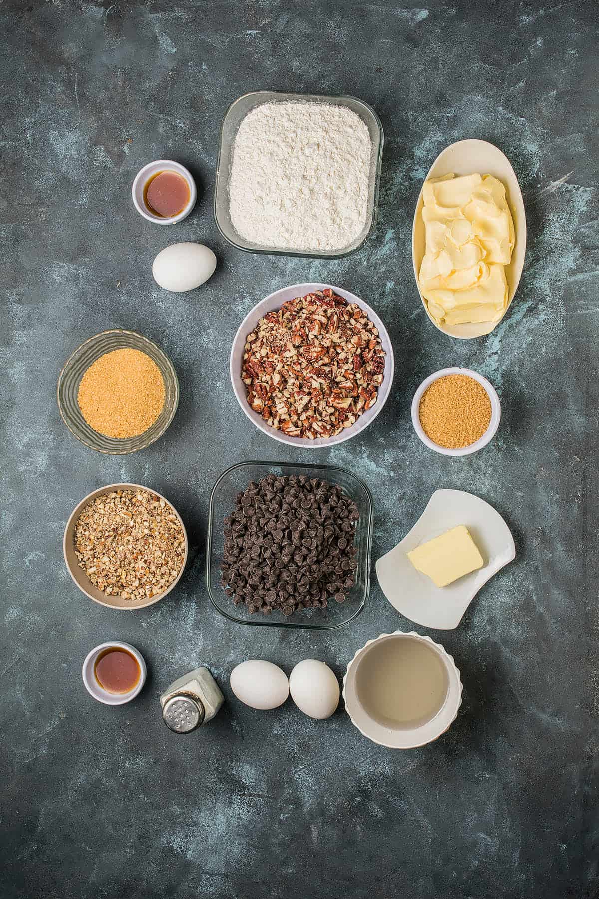 Several baking ingredients are arranged on a dark surface, ready for Dutch Oven Bread, including bowls of flour, chocolate chips, chopped nuts, sugar, cornmeal, butter, vanilla, and three eggs.