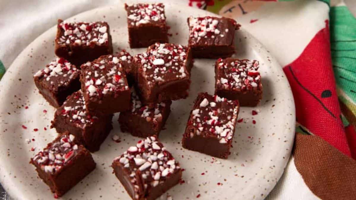 A plate of chocolate fudge squares topped with crushed peppermint candies, arranged on a white speckled dish with festive holiday-themed fabric underneath.