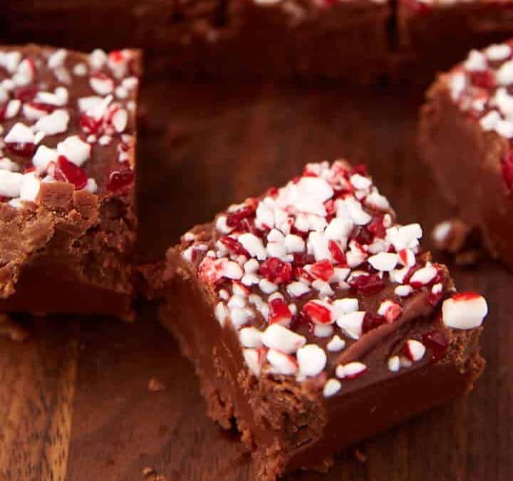 Squares of chocolate fudge topped with crushed peppermint candy, classic holiday treats you only see in December, are arranged on a wooden surface, with a few pieces separated from the main group in the foreground.