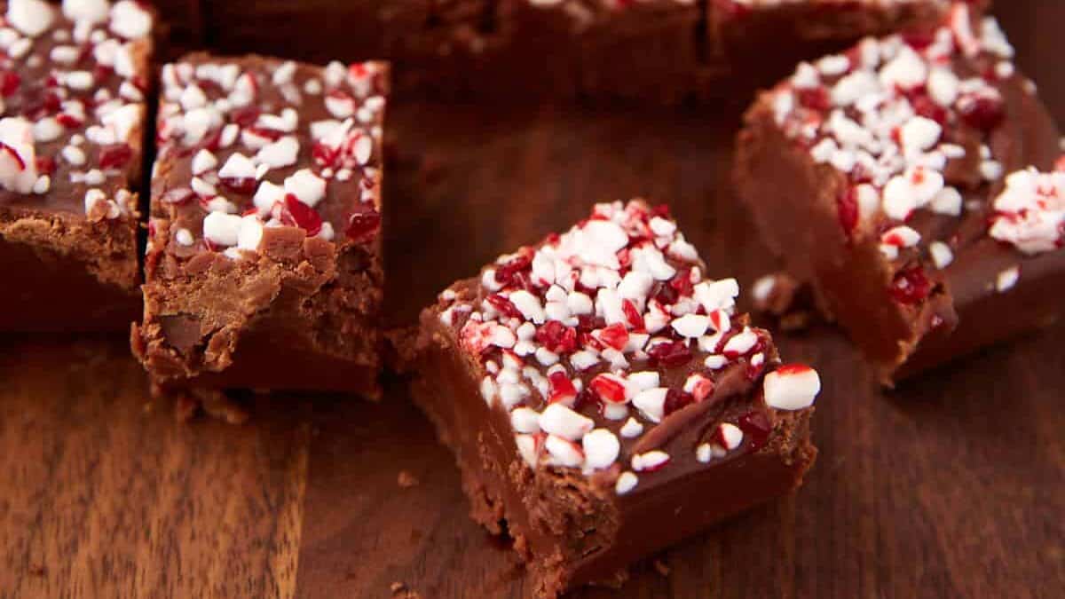Squares of chocolate fudge topped with crushed peppermint candy, classic holiday treats you only see in December, are arranged on a wooden surface, with a few pieces separated from the main group in the foreground.