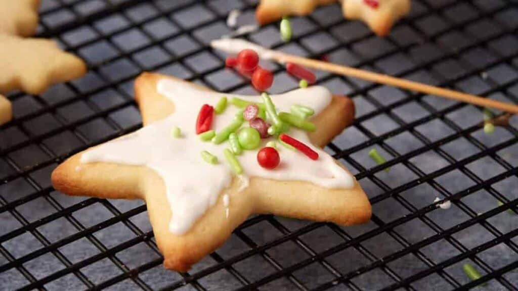 A star-shaped sugar cookie with white icing and red and green sprinkles sits on a cooling rack, capturing the spirit of holiday cookies getting baked nonstop, with a toothpick nearby.