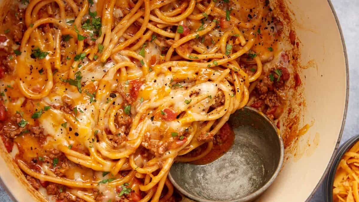 A pot of cheesy taco spaghetti casserole with meat sauce, topped with melted cheese and chopped parsley, with a metal ladle resting on the side.