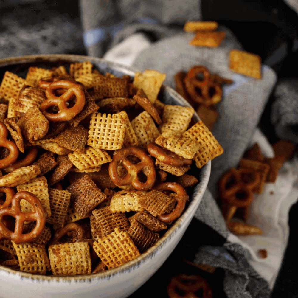 A close-up of a bowl filled with homemade snack mix, including square cereal pieces and pretzels, captures the essence of Southern Comfort Food, with more snack mix scattered on a gray cloth in the background.