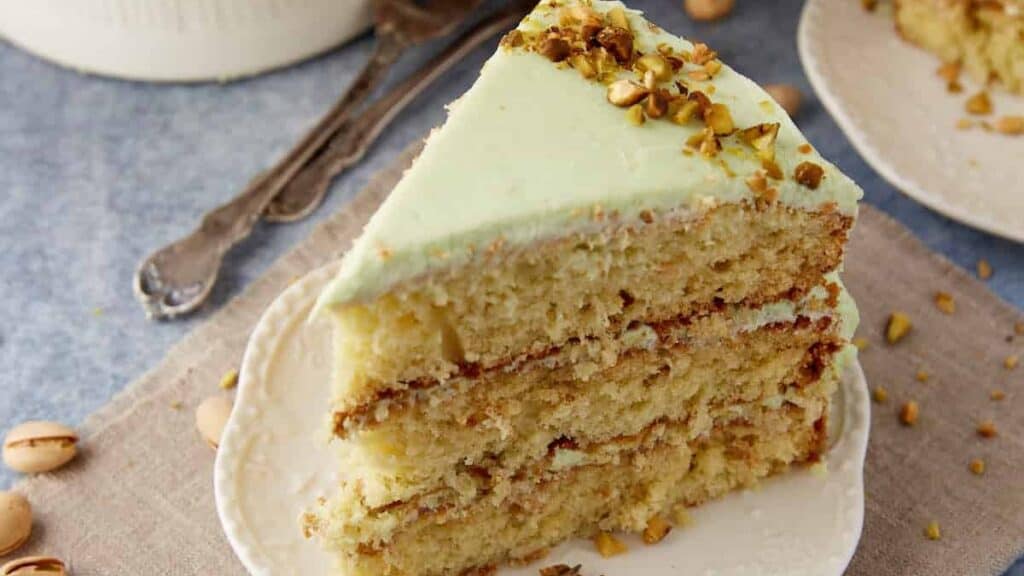 A slice of three-layer pistachio cake with light green frosting and chopped pistachios on top, served on a white plate—this festive dessert is perfect for christmas treats, bringing back old-fashioned holiday magic to your table.