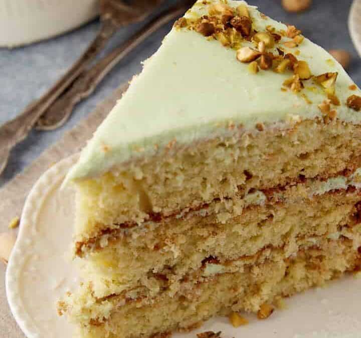 A slice of three-layer pistachio cake with light green frosting and chopped pistachios on top, served on a white plate—this festive dessert is perfect for christmas treats, bringing back old-fashioned holiday magic to your table.