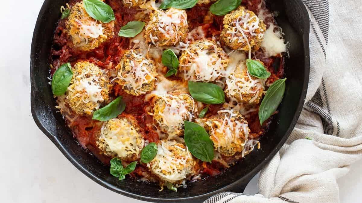 A cast iron skillet filled with baked meatballs in tomato sauce—an old school comfort food with a quiet update—topped with melted cheese and fresh basil, sits on a white marble surface, a beige striped towel wrapped around the handle.