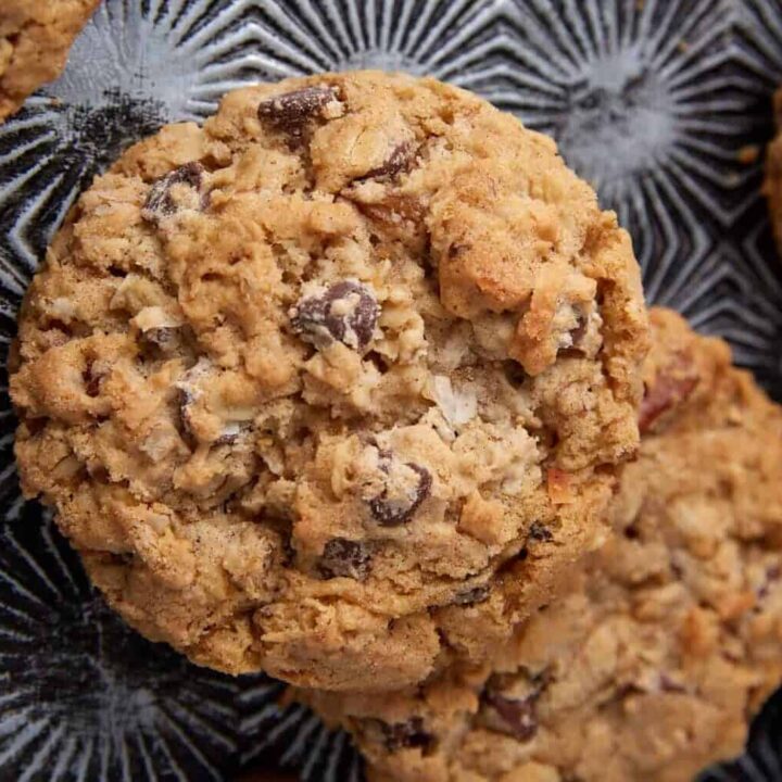 Close-up of oatmeal chocolate chip cookies on a textured dark surface, with chocolate chips scattered around. These chunky, golden brown treats are classic holiday cookies that always win the cookie swap.
