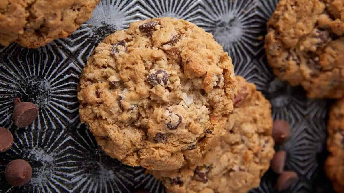 Close-up of oatmeal chocolate chip cookies on a textured dark surface, with chocolate chips scattered around. These chunky, golden brown treats are classic holiday cookies that always win the cookie swap.