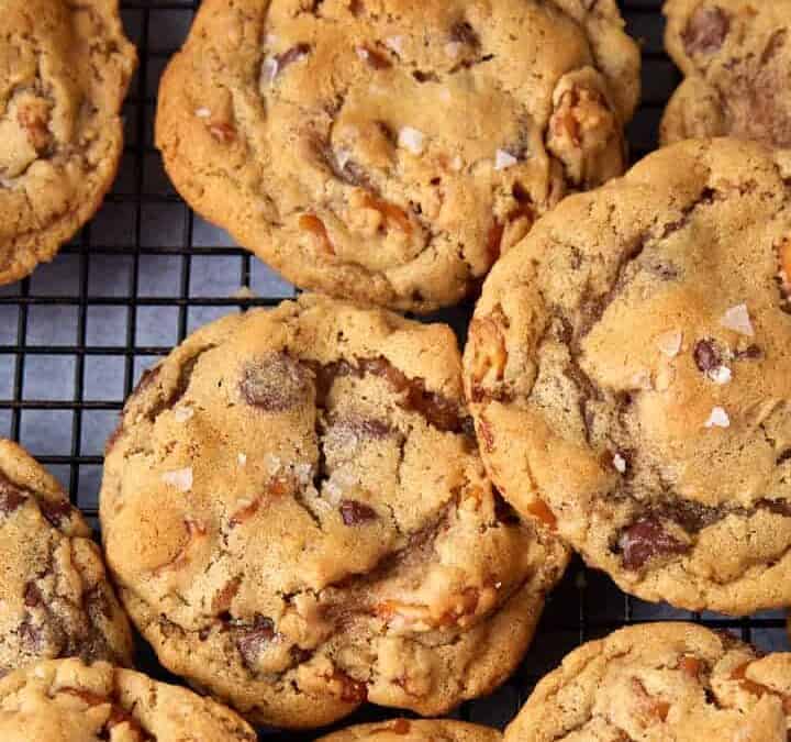 A batch of golden brown chocolate chip cookies with nuts—one of those classic Christmas cookies people never stop repeating—cooling on a black wire rack, each topped with a sprinkle of sea salt.