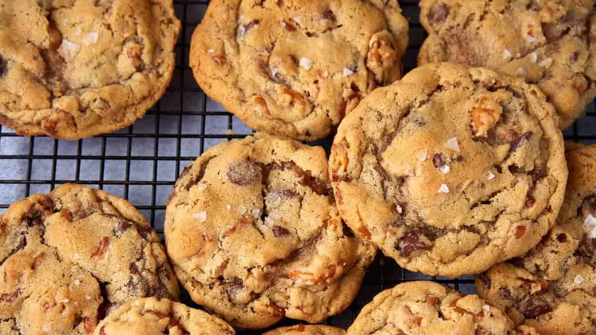 A batch of golden brown chocolate chip cookies with nuts—one of those classic Christmas cookies people never stop repeating—cooling on a black wire rack, each topped with a sprinkle of sea salt.