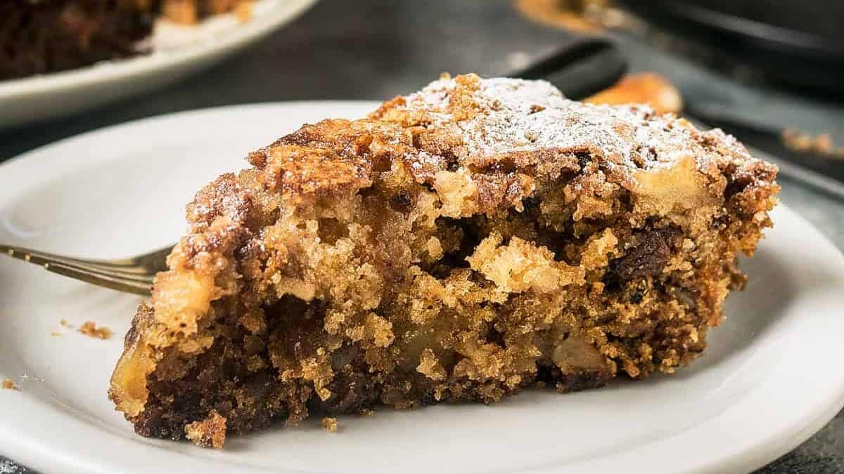 A close-up of an Apple Cinnamon Cake with a crispy, crumbly topping, served on a patterned plate. A 