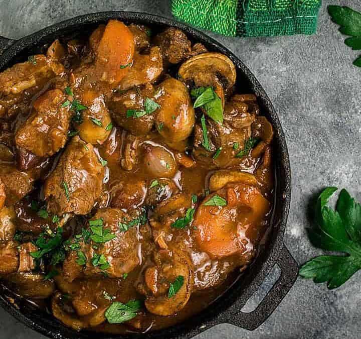 A bowl of hearty Beef Bourguignon with carrots and herbs sits on a gray surface near a spoon, a green towel, and fresh parsley, with a pot of more stew partially visible above.