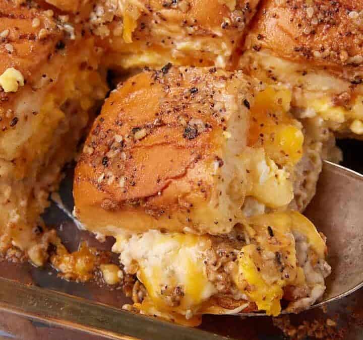 A glass baking dish filled with baked breakfast sliders made of golden brown rolls, scrambled eggs, and melted cheese, evokes the cozy comfort of a German apple pancake as a metal serving spoon lifts out one slider.