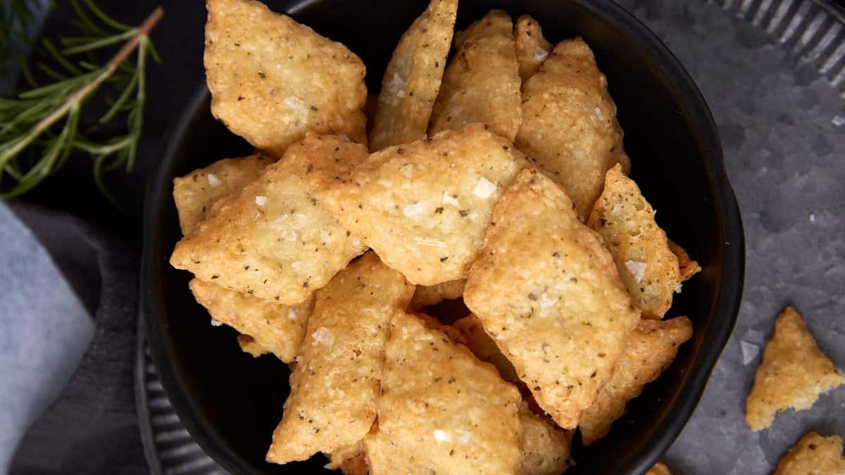 A black bowl filled with golden, crispy, diamond-shaped cheese crackers sprinkled with salt, placed on a metal tray with a sprig of rosemary in the background.