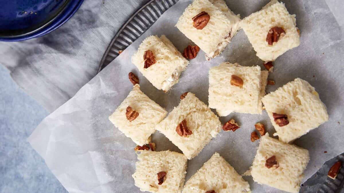 Squares of creamy white dessert topped with chopped pecans are arranged on parchment paper atop a metal tray. A dark blue plate and light grey napkin are partially visible nearby.
