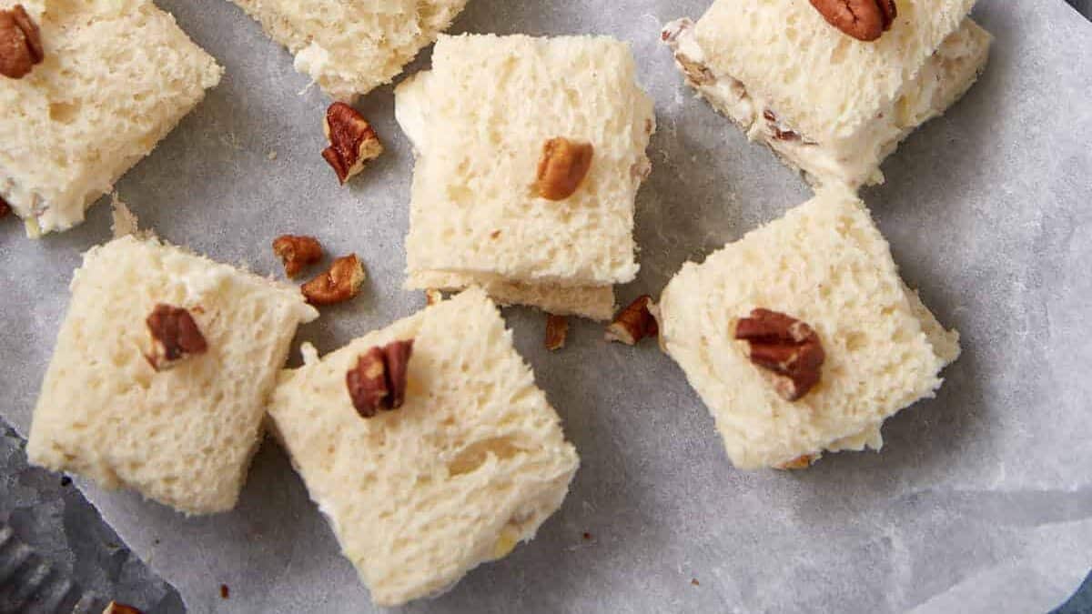 Squares of creamy white dessert topped with chopped pecans are arranged on parchment paper atop a me