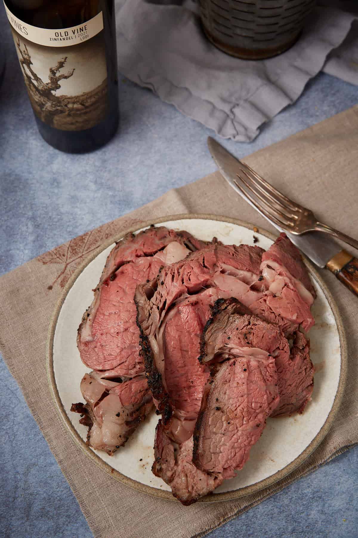 A plate with several slices of medium-rare standing rib roast sits on a beige napkin, next to a knife and fork. A bottle of wine and a folded gray napkin are visible in the background.