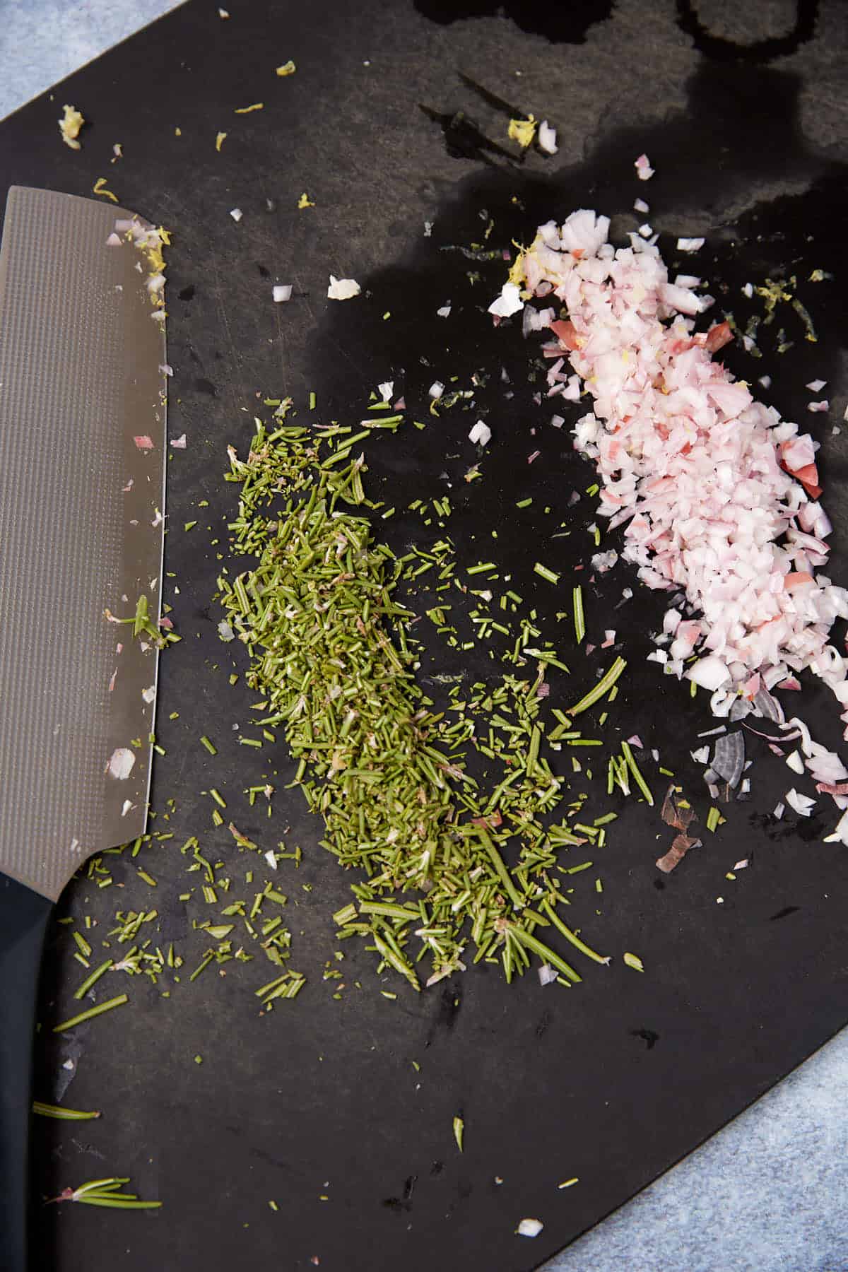 A close-up of a cutting board with a pile of chopped shallots and rosemary next to a kitchen knife, ready to season a standing rib roast.