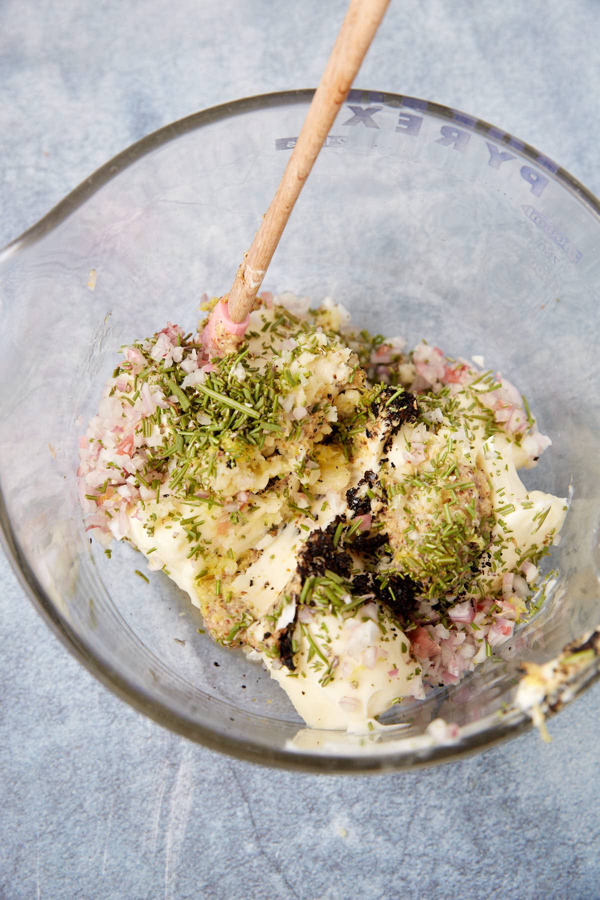 A glass mixing bowl contains chopped shallots, butter, dried herbs, and seasoning for a standing rib roast, with a wooden spoon resting inside. The ingredients are ready to be mixed.