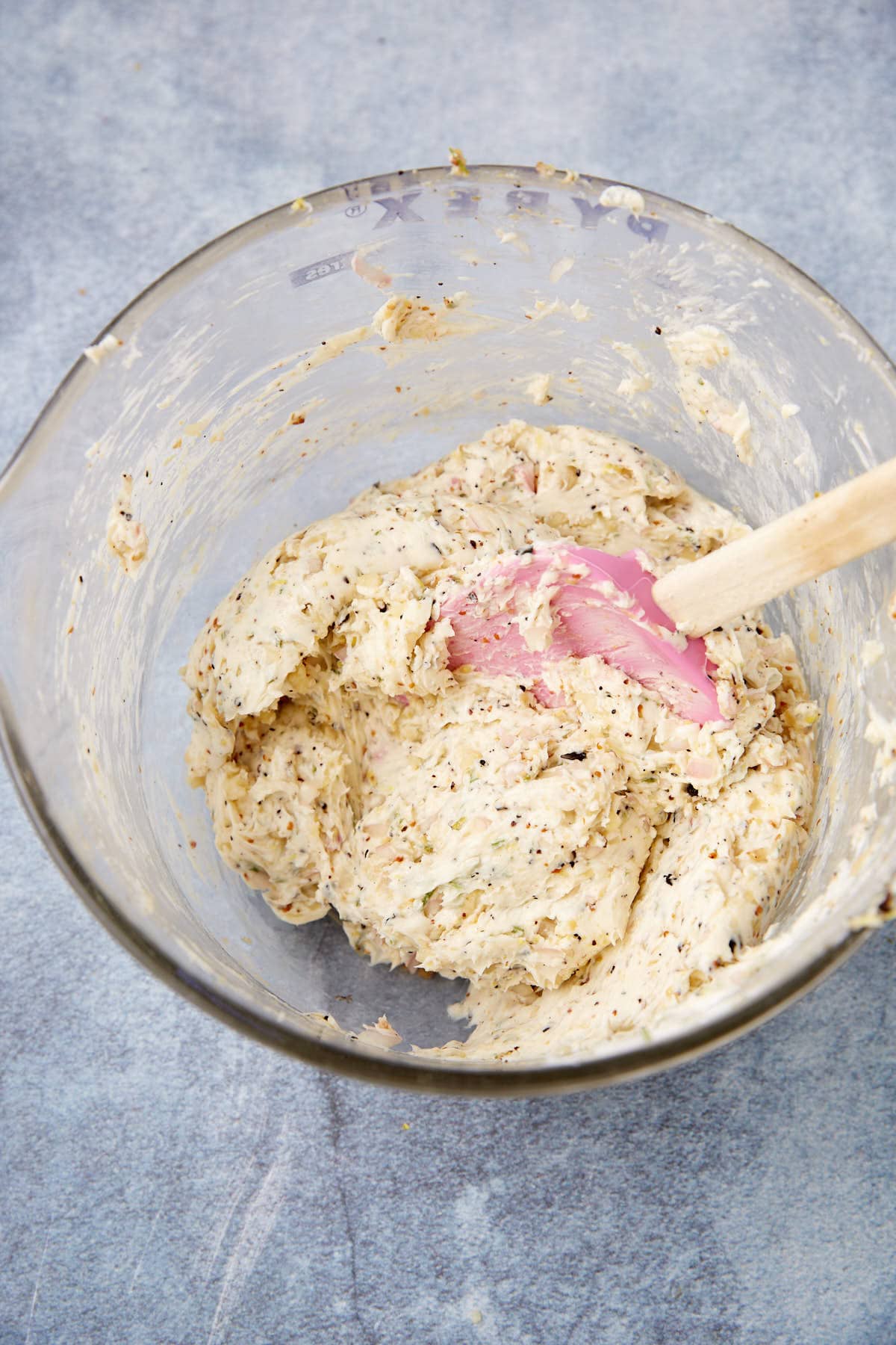 A glass bowl filled with mixed dough containing herbs and seasonings sits on a gray textured surface. A pink spatula with a wooden handle rests in the dough, ready to complement your standing rib roast.