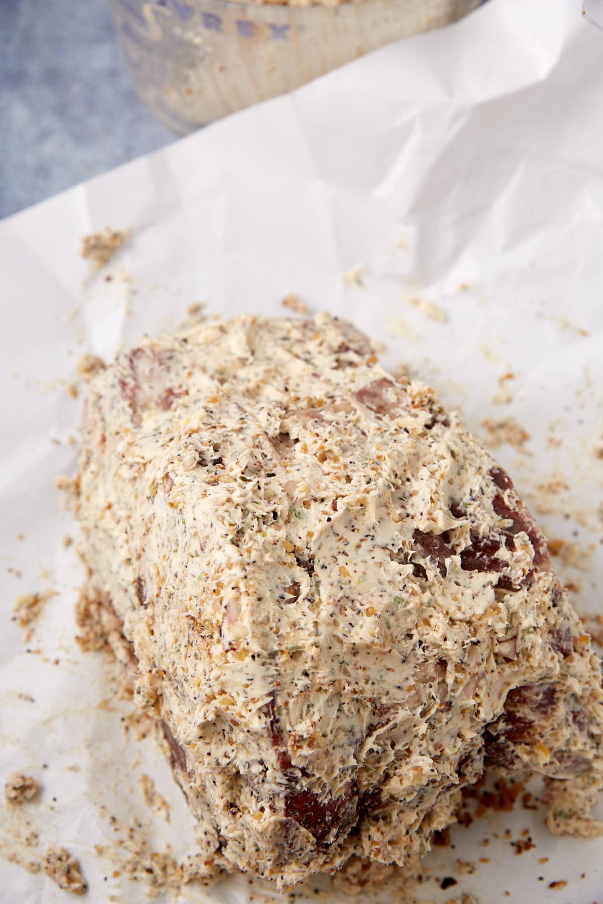 A standing rib roast coated with a thick layer of seasoned herb and spice butter sits on a sheet of white parchment paper, ready to be cooked.