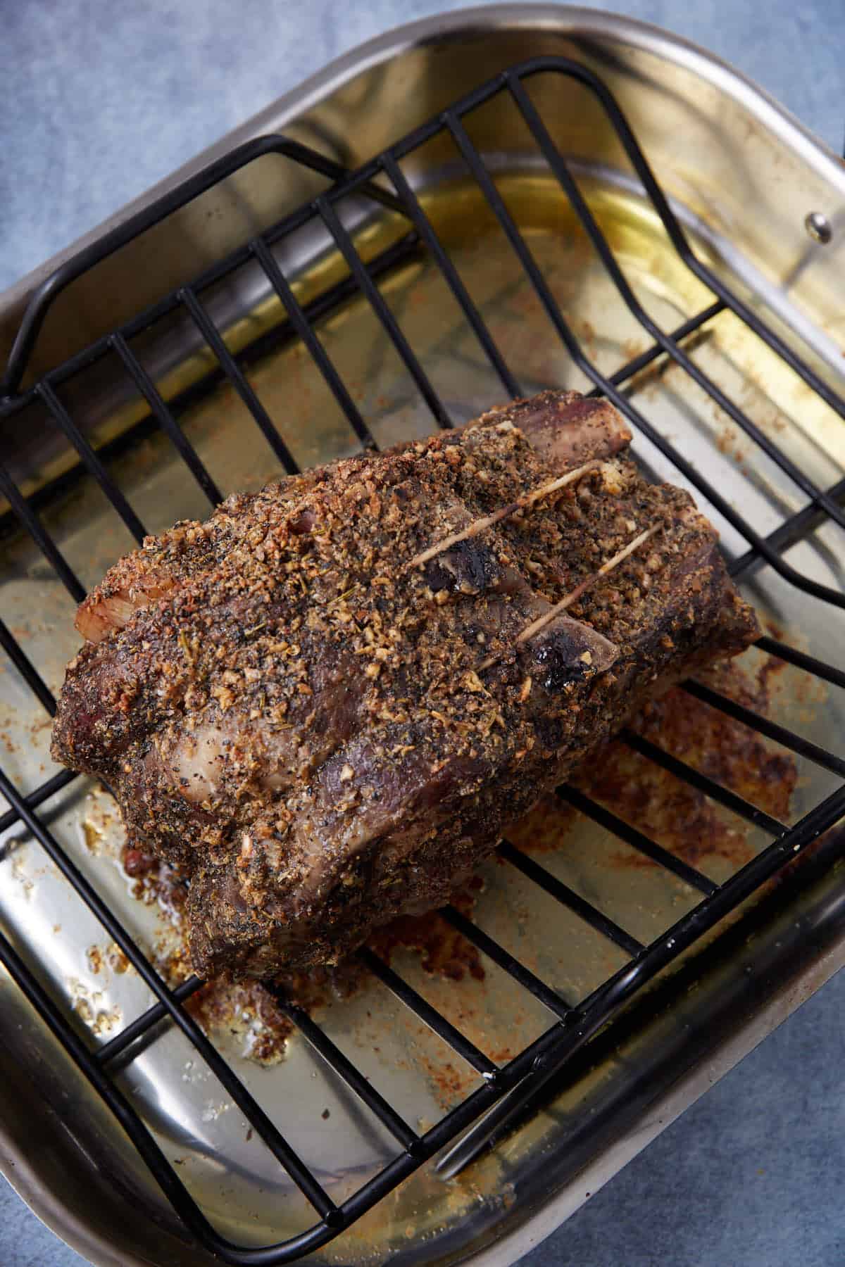 A cooked, seasoned standing rib roast is resting on a black roasting rack inside a metal roasting pan, with juices collected at the bottom of the pan.