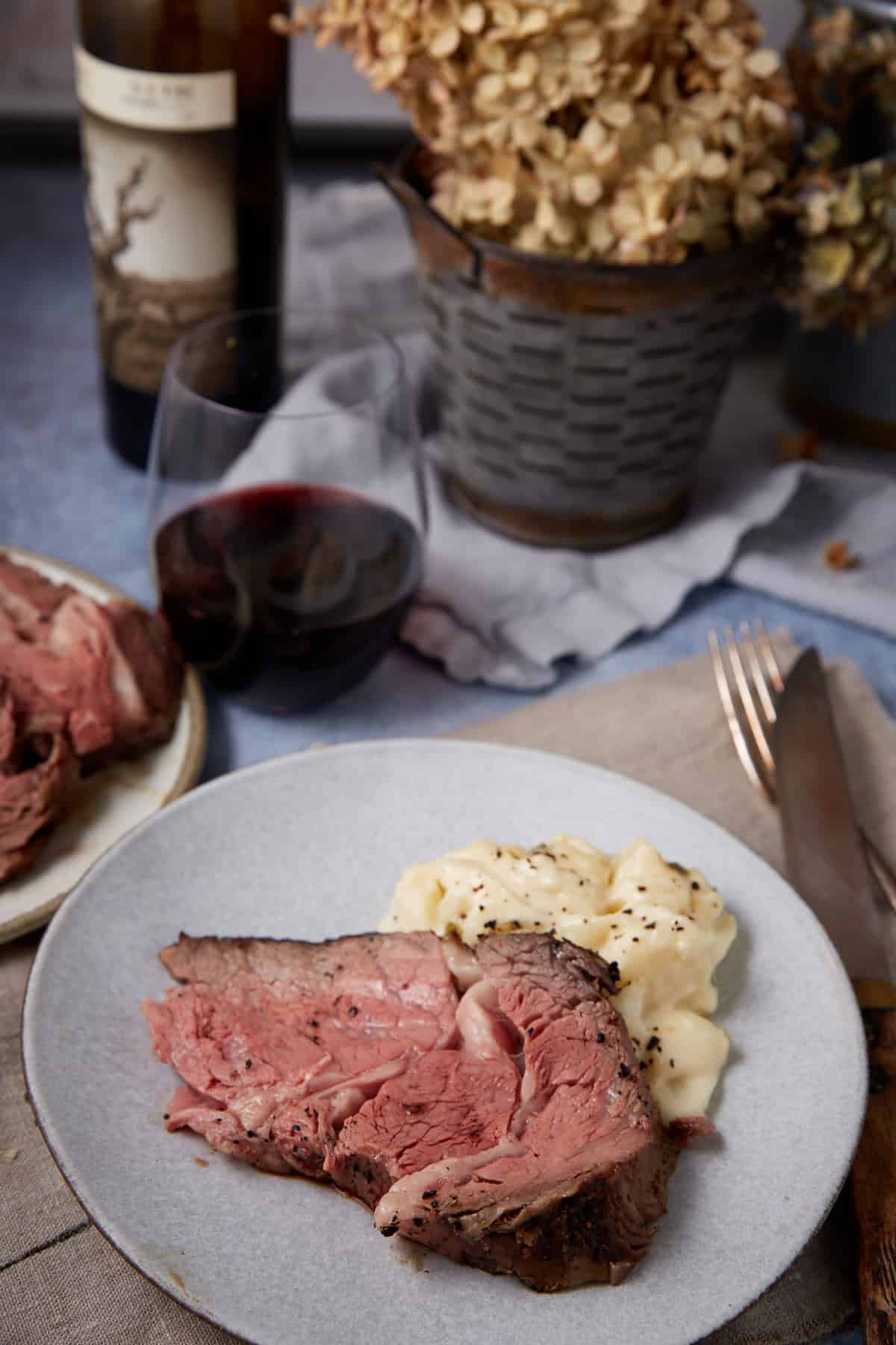 A plate of sliced standing rib roast and creamy mashed potatoes sits on a table with a fork and knife. A glass of red wine, a bottle, and a rustic pot of dried flowers are in the background.
