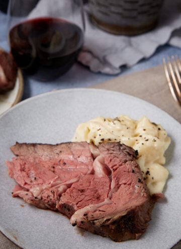 A plate with a thick slice of medium-rare standing rib roast and a serving of mashed potatoes, sprinkled with black pepper. In the background, there is a glass of red wine and cutlery on a napkin.